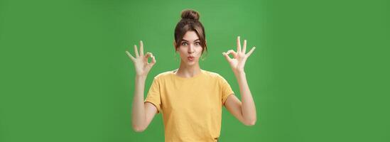 portrait de idiot mignonne et magnifique caucasien femme avec peigné cheveux dans rond des boucles d'oreilles et Jaune T-shirt montrant d'accord, non problème geste pliant lèvres, réaffirmant patron emploi va bien plus de vert mur photo