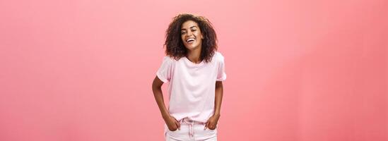 éclairer en haut la vie avec sourire. insouciant charismatique content africain américain femelle avec afro coiffure dans branché tenue en riant en dehors bruyant de joie et amusement en portant mains dans les poches plus de rose mur photo