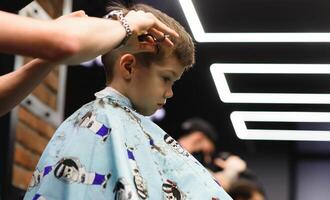 côté vue de mignonne peu garçon avoir la Coupe de cheveux par coiffeur à le salon de coiffure. photo