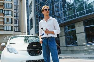 homme en portant Puissance mise en charge câble pour électrique voiture dans Extérieur voiture parc. et il s Aller à relier le voiture à le mise en charge station dans le parking lot près le achats centre photo