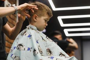 côté vue de mignonne peu garçon avoir la Coupe de cheveux par coiffeur à le salon de coiffure. photo