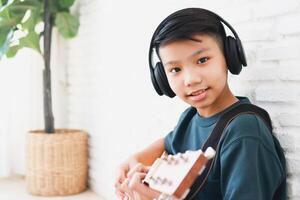 asiatique garçon portant écouteurs avec noir la musique asseoir et jouer guitare dans le maison. la musique apprentissage concept, la musique compétence formation photo