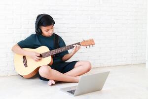 asiatique garçon portant noir la musique écouteurs en jouant guitare dans le maison apprendre la musique en ligne par une portable. la musique apprentissage concept, la musique compétence entraînement. copie espace photo