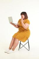 asiatique graisse femme séance sur une chaise sur une blanc Contexte elle détient une portable à travail. concept de travail en ligne. mode robe photo