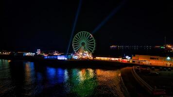 la nuit vue de une allumé ferris roue par le l'eau avec coloré reflets et projecteurs dans le ciel dans backpool, Angleterre. photo