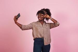 jolie femme afro-américaine en vêtements décontractés prend un selfie et montre un geste avec deux doigts sur fond rose dans le studio photo