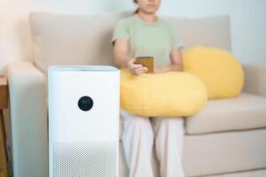 air purificateur avec femme se détendre et utilisation téléphone intelligent sur canapé. purification système pour filtre et nettoyage poussière pm2,5 hepa et virus dans maison. allergie, pur air, santé, bien-être mode de vie et air la pollution photo