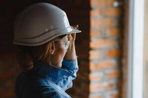 attrayant femelle construction ouvrier dans casque. sur de soi Jeune spécialiste dans à carreaux bleu chemise dans jeans permanent dans vide chambre. intérieur conception et rénovation un service photo