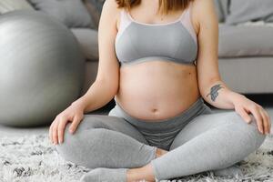 fermer coup de méconnaissable Enceinte femme méditer à maison, attendant Dame séance dans lotus position avec étreint mains, pratiquant yoga pour en bonne santé grossesse, tondu image avec gratuit espace photo