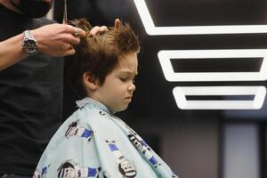 côté vue de mignonne peu garçon avoir la Coupe de cheveux par coiffeur à le salon de coiffure. photo