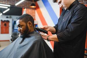 portrait de Jeune noir homme étant taillé avec professionnel électrique tondeuse machine dans salon de coiffure.male beauté traitement concept. Jeune africain gars avoir Nouveau la Coupe de cheveux dans coiffeur salon photo