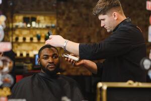 fermer de processus de garniture de cheveux dans coiffeur magasin. qualifié coiffeur en gardant tondeuse dans mains et corriger forme de cheveux à Masculin client séance sur chaise. concept de la Coupe de cheveux et rasage. photo