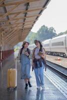 deux Jeune asiatique copains les filles avec sacs à dos à chemin de fer station attendre pour former, deux magnifique femmes en marchant le long de Plate-forme à train station photo