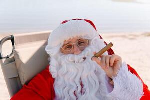 Père Noël dans des lunettes fume une cigare tandis que mensonge sur une Soleil fainéant sur une Lac plage photo