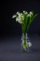 une bouquet de fleurs de lys de le vallée sur une noir Contexte avec endroit pour texte. délicat blanc printemps fleurs. bouquet dans une verre bouteille. espace de copie. photo