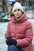 une caucasien femme dans une tricoté chapeau et hiver vers le bas veste détient une papier tasse de café dans sa mains. photo