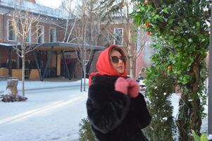 une Jeune femme, une fille dans des lunettes de soleil, une rouge écharpe et une foncé fourrure manteau, des stands près une vert arbre à le entrée à une ville café. hiver saison. photo