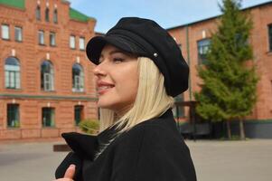 portrait de une caucasien blond fille dans une noir manteau et casquette en marchant le long de une ville rue. photo