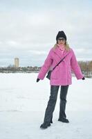 une caucasien blond fille dans une brillant rose hiver fourrure manteau, une noir chapeau, cuir pantalon et des lunettes de soleil des stands dans le neige dans une parc dans l'hiver. photo