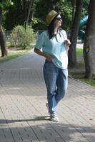 une Jeune femme dans une été paille chapeau, des lunettes de soleil, une lumière colorée polo chemise et jeans, des stands sur le route et regards une façon dans le distance. photo