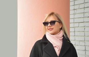 Urbain portrait de une moderne caucasien blond fille dans une noir manteau, rose écharpe et des lunettes de soleil. photo