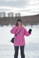 à la mode caucasien fille dans une chaud rose hiver faux fourrure manteau, noir chapeau et des lunettes de soleil, parlant sur une mobile téléphone contre le toile de fond de neige dans une parc dans l'hiver. photo