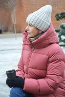 portrait de une mignonne caucasien femme dans chaud hiver vêtements, à la recherche à le côté et en portant une papier verre de café dans sa mains. photo