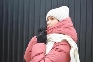 une Jeune femme dans hiver vêtements - une vers le bas veste, une tricoté chapeau, gants et une lumière écharpe sur une foncé Contexte. portrait de une femme sur une hiver journée. photo