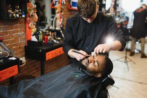 côté vue de sérieux homme avec élégant moderne la Coupe de cheveux à la recherche vers l'avant dans coiffeur magasin. main de coiffeur en gardant tout droit le rasoir et Coupe branché rayures sur tête de client. photo