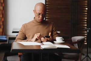 africain homme d'affaire parlant sur téléphone séance à café tableau, occupé entrepreneur travail lointain dans café maison avec portable papiers Parlant sur mobile, noir homme fabrication appel ayant le déjeuner dans cafétéria. photo