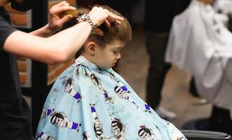 une peu mignonne garçon est assis dans une salon de coiffure à le styliste, une écolier est avoir cheveux Couper dans une beauté salon, une enfant à une chez le coiffeur, une court Pour des hommes la Coupe de cheveux photo