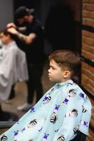 côté vue de mignonne peu garçon avoir la Coupe de cheveux par coiffeur à le salon de coiffure. photo