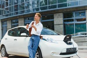 homme mise en charge le sien luxe électrique voiture à Extérieur station dans de face de moderne Nouveau ville bâtiments photo