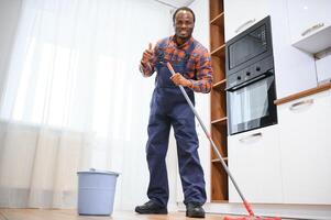 Jeune africain homme lavages le sol avec une balai dans le pièce photo