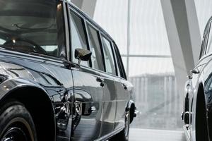 Vue du côté conducteur limousine classe affaires rétro noir avec miroir chromé garé près d'une autre voiture d'époque dans un bâtiment léger photo