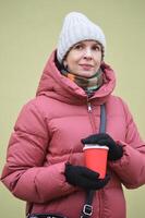 portrait de une caucasien femme avec une hiver vers le bas veste et une tricoté chapeau avec une rouge papier tasse de café dans sa mains. photo