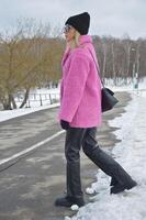 une Jeune magnifique femme dans une élégant rose fourrure manteau, noir chapeau et pantalon des promenades le long de une hiver route dans une ville parc photo