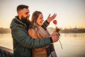 content homme donnant rouge Rose à le sien femme tandis que elles ou ils prendre plaisir dépenses temps ensemble sur une le coucher du soleil. photo