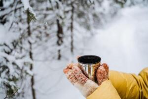 thermo agresser dans main. une main détient une agresser de chaud thé. boisson thé dans le hiver dehors. une métal tasse avec une boisson est tenue par une personne. thé de une thermos. photo