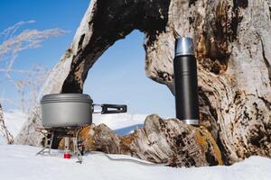 touristique vaisselle fermer. une pot de nourriture des stands sur une gaz brûleur, une noir thermos avec thé, une hiver randonnée, neige dans le forêt, cuisine dans la nature. photo