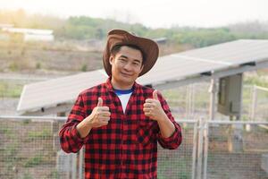 Beau asiatique homme porte chapeau, rouge plaid chemise, supporter à côté de solaire panneau à agriculture atterrir. les pouces en haut. concept, agriculteur installée photovoltaïque pour utilisation dans agriculture pour électricité. photo