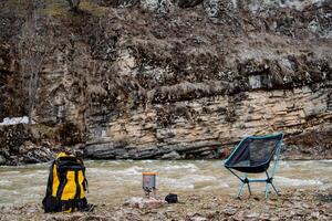 camping sur le rivière banque, une Jaune sac à dos avec randonnée équipement, une pot sur une gaz brûleur, une pliant chaise pour repos, une camping lieu. photo