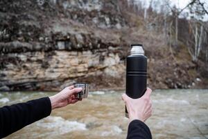 une touristique la personne détient dans le sien mains une thermos avec une tasse contre le Contexte de le rivière, le concept de vaisselle pour chaud les boissons dans une une randonnée dans le montagnes, printemps touristique vaisselle photo