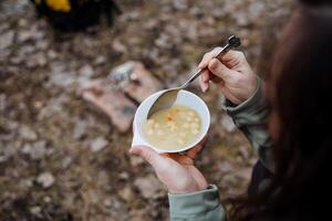 le fille détient dans sa mains une assiette de soupe dans une une randonnée dans nature, une touristes petit-déjeuner, en mangeant liquide nourriture avec une cuillère dans le forêt, le déjeuner dans le printemps dans le forêt. photo