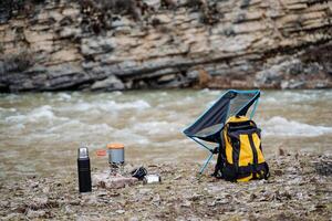 touristique camp sur le banques de une Montagne rivière, Jaune sac à dos des stands sur le sol, pliant ultra-léger chaise, camping ustensiles, le déjeuner dans nature, thermos avec thé, pot photo