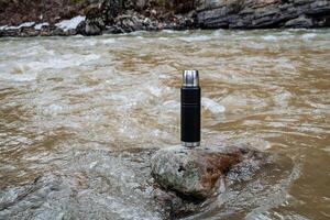 le thermos des stands sur une pierre dans le l'eau. vide thermos pour thé, bouteille pour chaud boissons. camping ustensiles, touristique équipement. photo