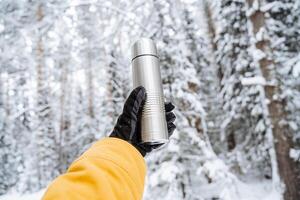 main avec une thermos contre le Contexte de le forêt dans l'hiver. acier thermos. métal thermos. couvert de neige blanc forêt, taïga dans le neige. tendu bras. photo