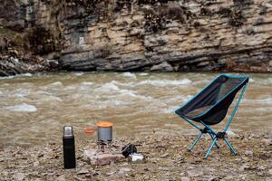 touristique parking près le rivière, camping sur le rive de le réservoir, cuisine pour cuisine sur une randonnée, pliant chaise, thermos avec thé, pot, vaisselle sur le sol. photo