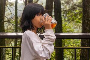 un asiatique peu fille permanent et en buvant une Plastique tasse de la glace thé sur le balcon avec la nature ou des arbres Contexte photo