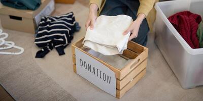 don, asiatique Jeune femme séance pack objet à maison, en mettant sur des trucs dans faire un don boîte avec seconde main vêtements, charité portion et nécessiteux personnes. réutilisation recycler photo